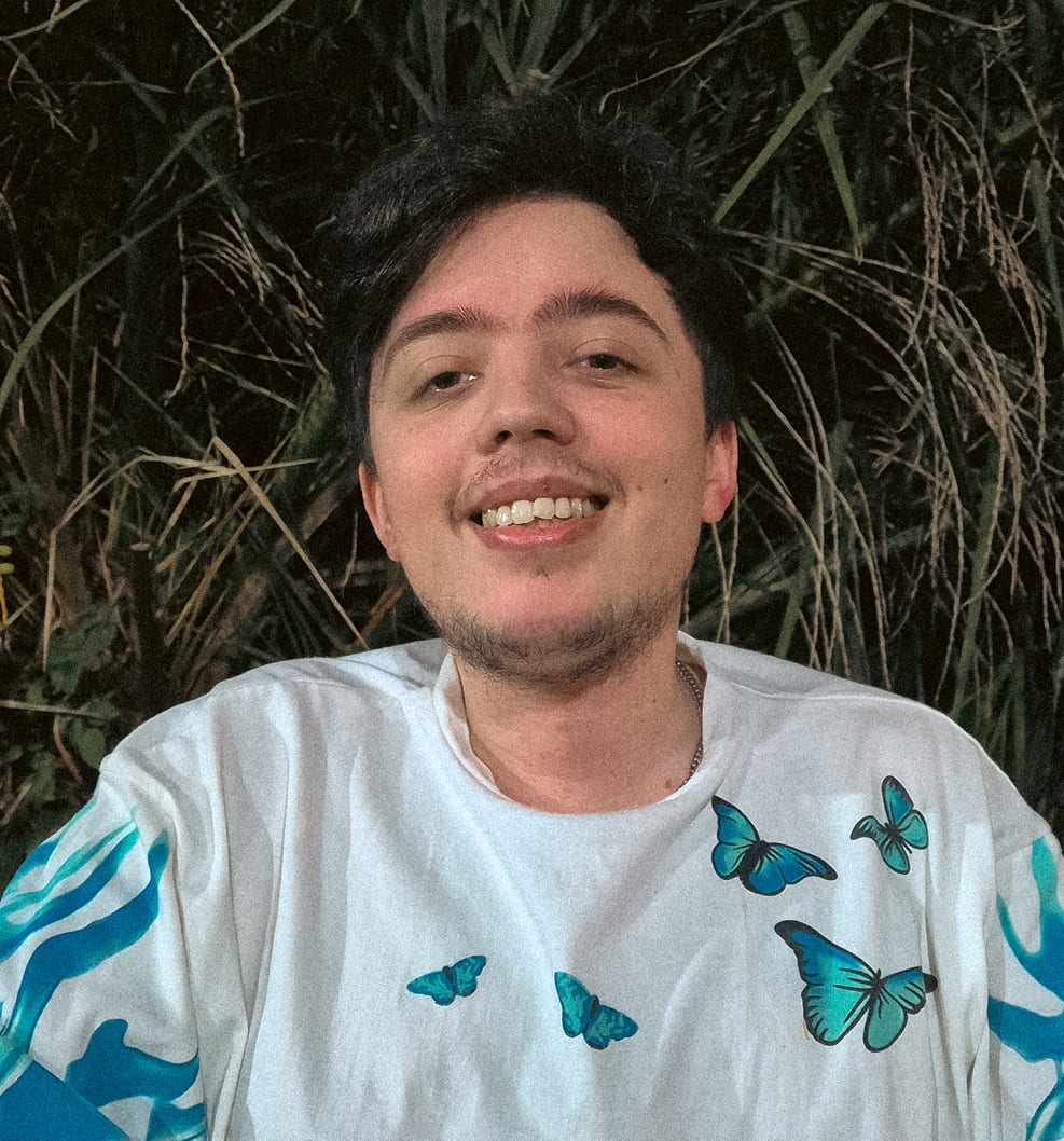 Agustin sonriendo, con remera blanca con mariposas celestes, frente a pastos altos.